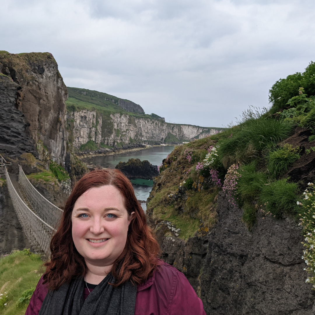 Lauren, a white woman with red hair and blue eyes, stands in front of the Carrick-a-Rede bridge in Northern Ireland